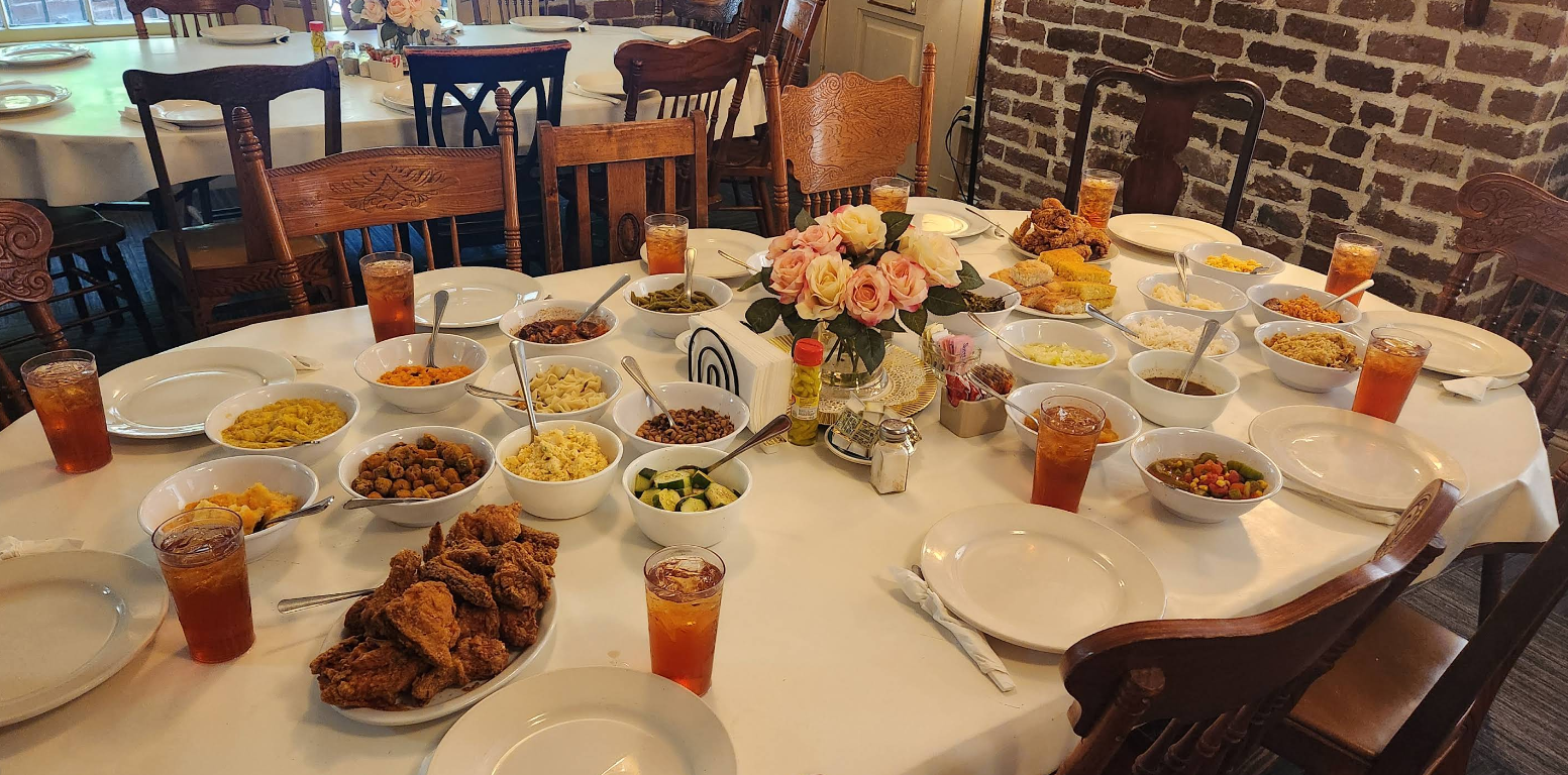 A round table set for a meal with plates, glasses of iced tea, fried chicken, mac n cheese and other side dishes in bowls, plus a floral centerpiece, surrounded by wooden chairs in a cozy dining room.