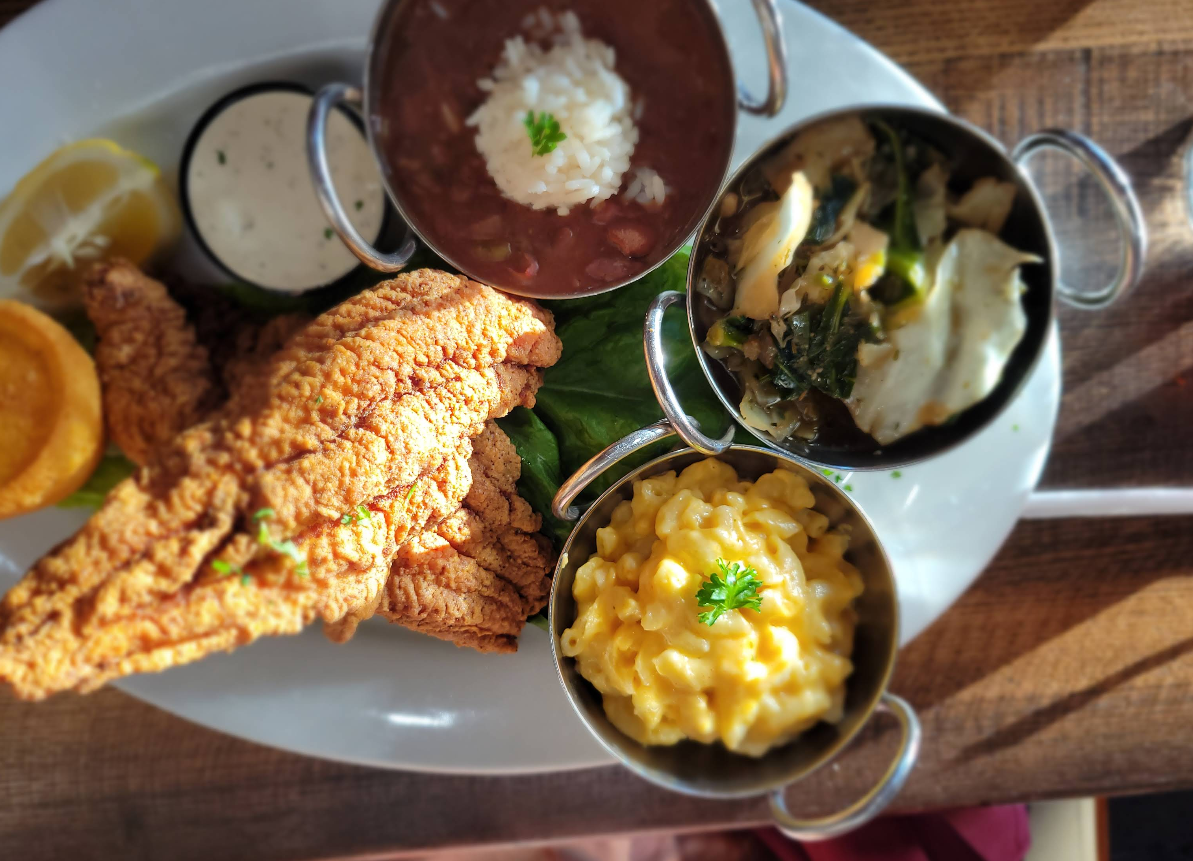 A plate with fried catfish, lemon slices, dipping sauce, creamy mac n cheese, a bowl of beans and rice, and cooked greens, all arranged in small metal bowls.