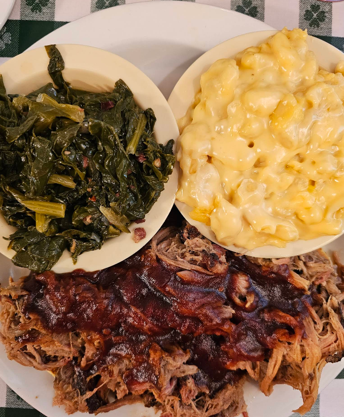 A plate of barbecue pulled pork with tangy barbecue sauce, served alongside creamy mac n cheese and a side of cooked collard greens in separate bowls, all set on a green and white checkered tablecloth.