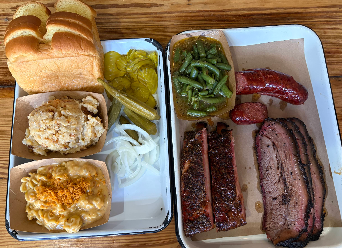 Two trays with barbecue food: sliced brisket, sausage, ribs, green beans, creamy mac n cheese, potato salad, sliced onions, pickles, pickled okra, and four thick slices of bread on a wooden table.