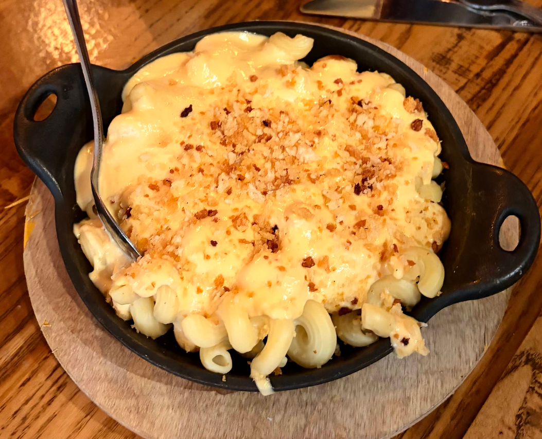 A black dish filled with creamy mac n cheese, topped with a golden breadcrumb crust, sits on a wooden table with a spoon inside, ready to serve.