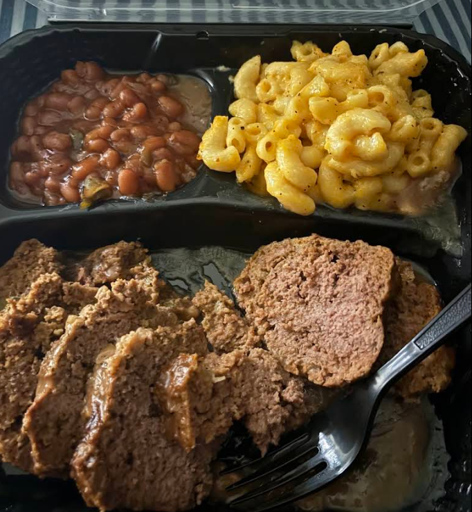 A black takeout container with three sections holds baked beans, creamy mac n cheese, and several slices of meatloaf. A plastic fork rests on the meatloaf.
