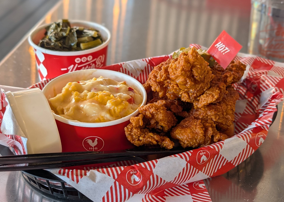 A basket of crispy fried chicken labeled "HOT!", served with creamy mac n cheese, collard greens in a cup, and wrapped utensils, all on red and white checkered paper.
