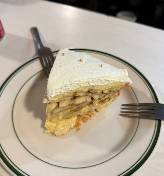 A slice of banana cream pie with a flaky crust, layered banana filling, and a thick topping of whipped cream sits on a white plate with a green rim, flanked by two forks—perfect alongside classic apple pies.