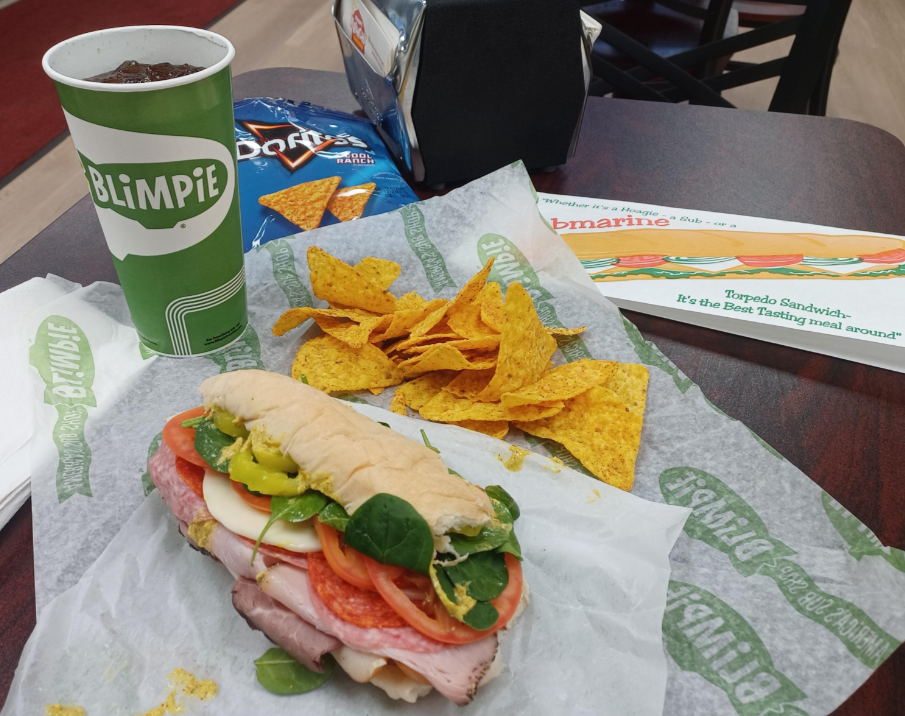 A deli sandwich from one of the top sandwich and sub chains features ham, pepperoni, cheese, tomato, spinach, and banana peppers on branded paper beside Doritos chips, a green Blimpie drink cup with a straw, and a napkin on a dark table.