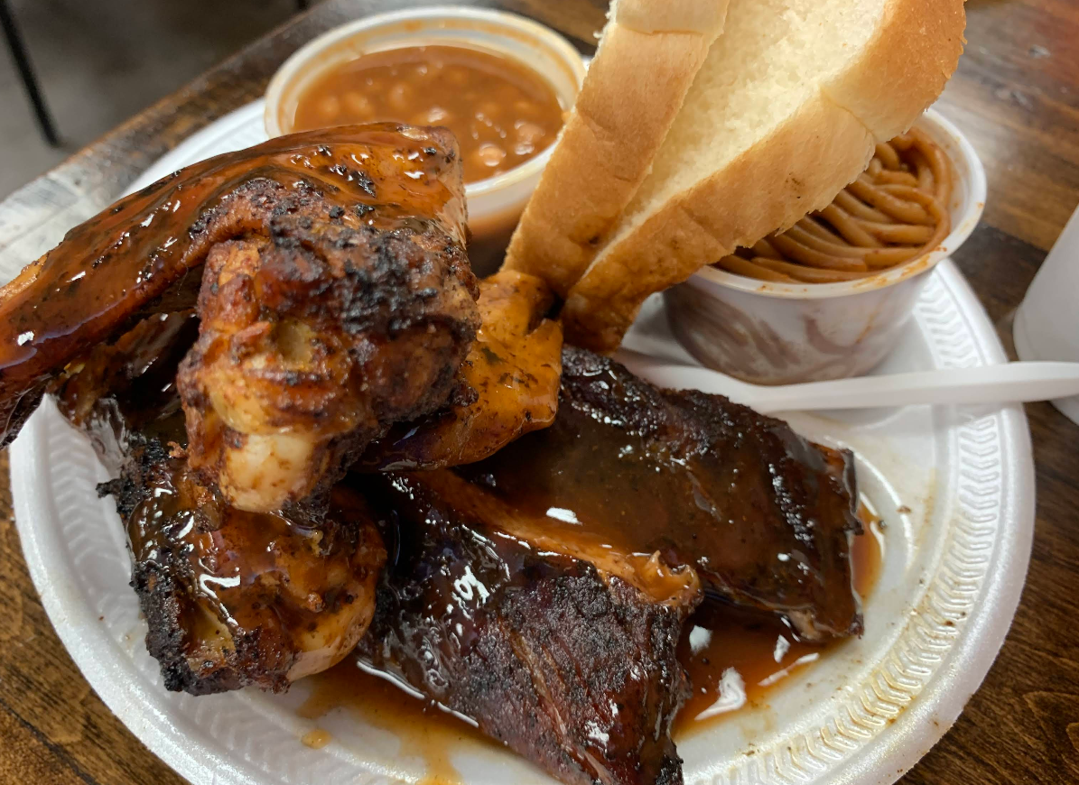 A plate of barbecue featuring the best ribs in America—glazed and tender—grilled chicken, slices of white bread, baked beans, and spaghetti, all served on a white disposable plate with a plastic fork.