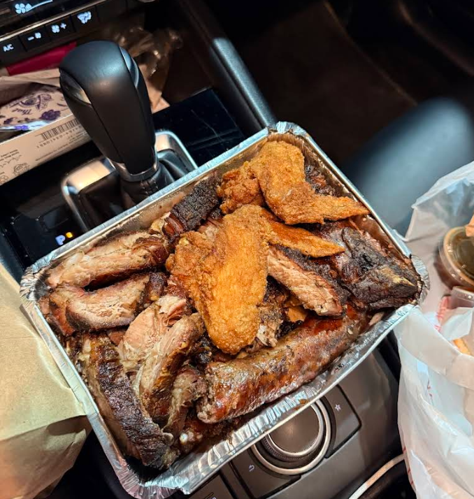 A foil tray filled with assorted barbecued meats and chicken wings—featuring some of the best ribs in America—sits on the center console of a car, surrounded by bags and a gear shift.