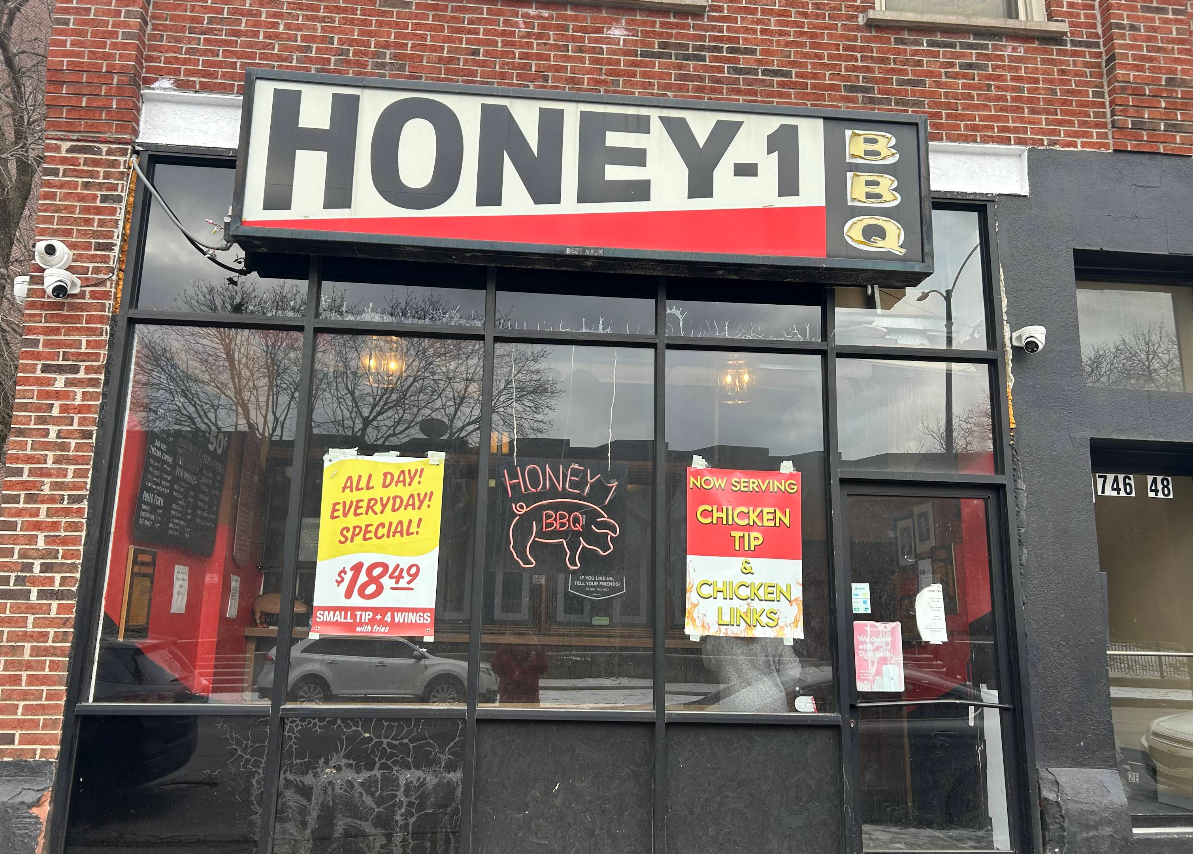 Storefront of Honey-1 BBQ with large sign above entrance, neon pig in the window, and posters advertising specials like chicken tip, small tip with 4 wings for $18.49, and chicken links—home of some of the best ribs in America. Brick building exterior.