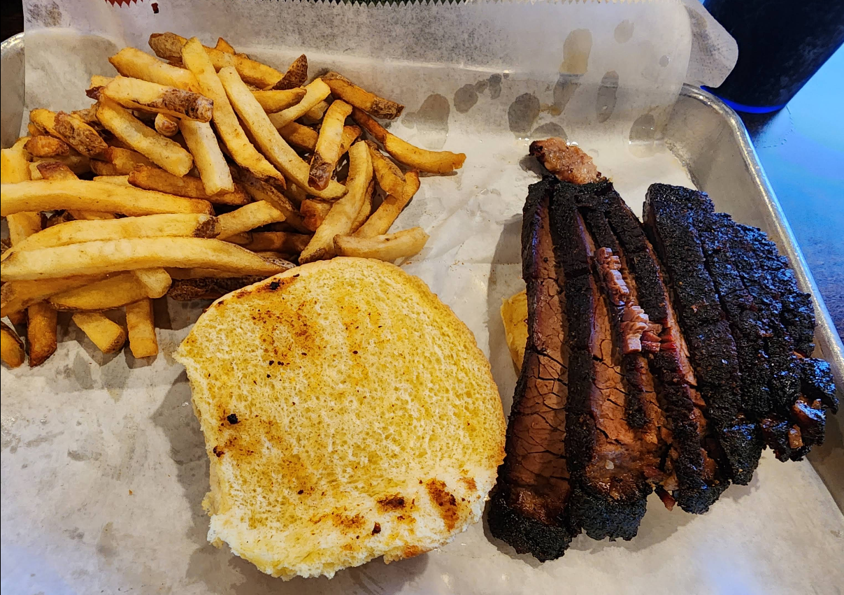 A food tray with a serving of French fries, a toasted sandwich bun, and several slices of smoked brisket with a dark bark crust—just what you’d expect at a spot known for some of the best ribs in America. A drink cup is partly visible in the background.