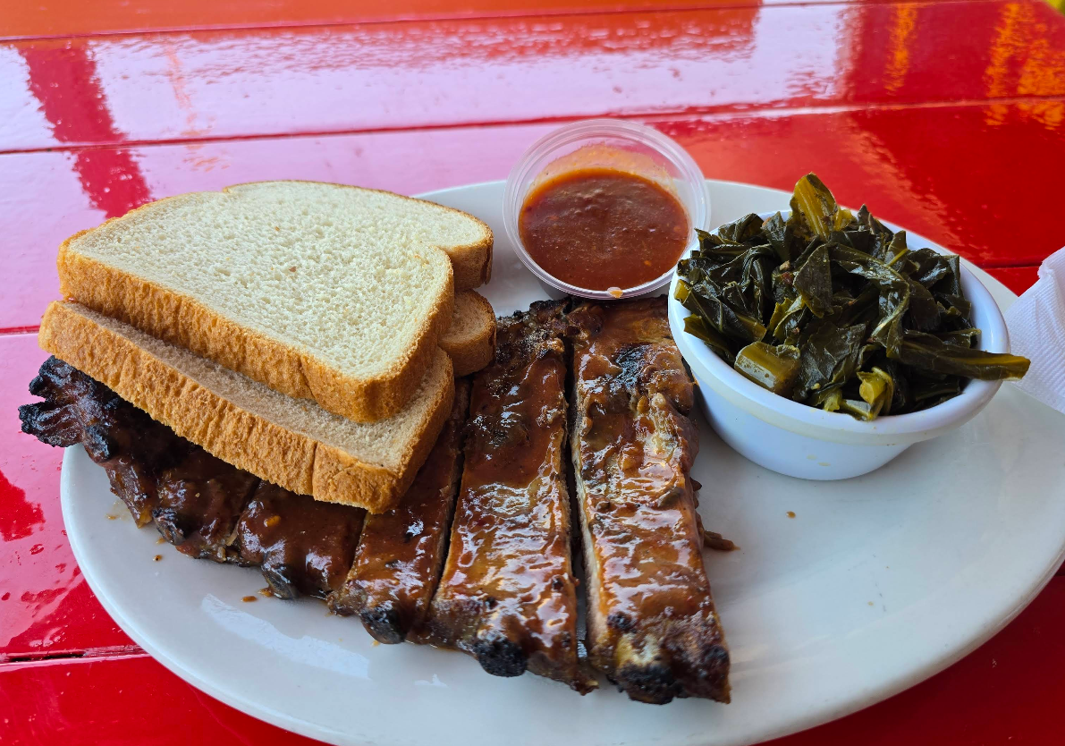 A plate with barbecue ribs covered in sauce—boasting some of the best ribs in America—served with three slices of white bread, a cup of barbecue sauce, and a bowl of cooked collard greens on a red table.