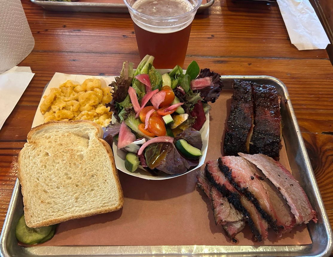 A tray with slices of white bread, macaroni and cheese, a green salad with tomatoes and pickled onions, the best ribs in America, smoked brisket slices, and a cup of amber beer on a wooden table.