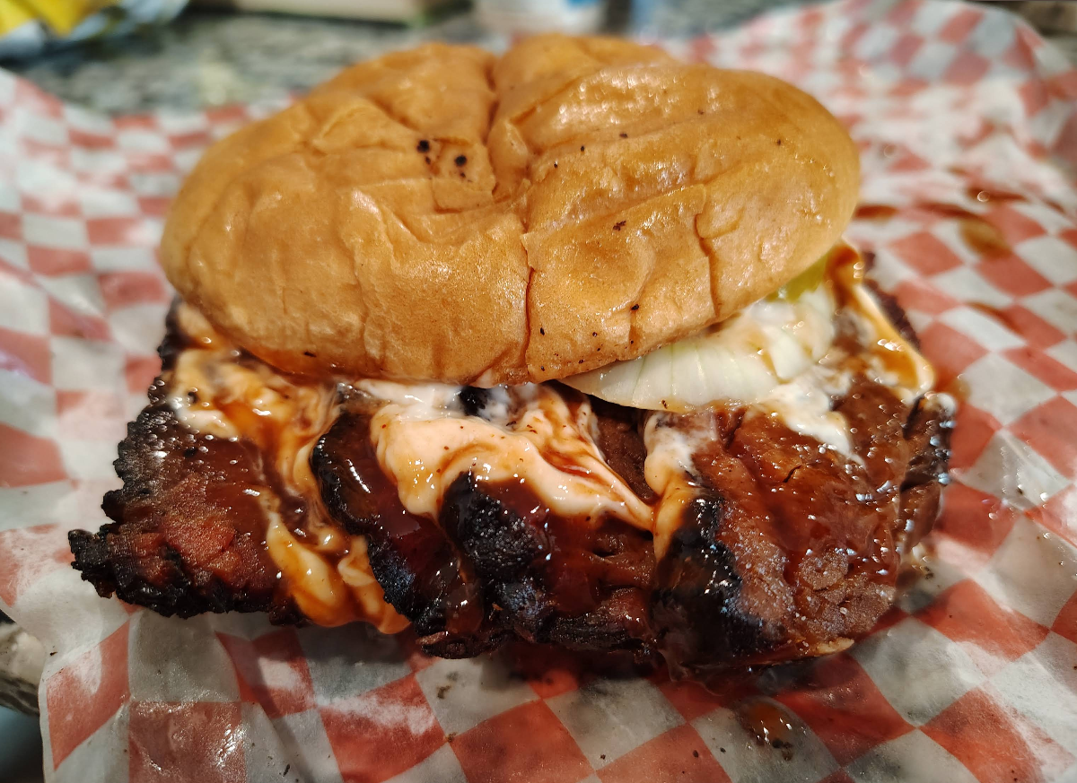 A close-up of a brisket sandwich with barbecued beef, sliced onions, and sauce on a soft bun, placed on red-and-white checkered paper—made at a spot famous for some of the best ribs in America.