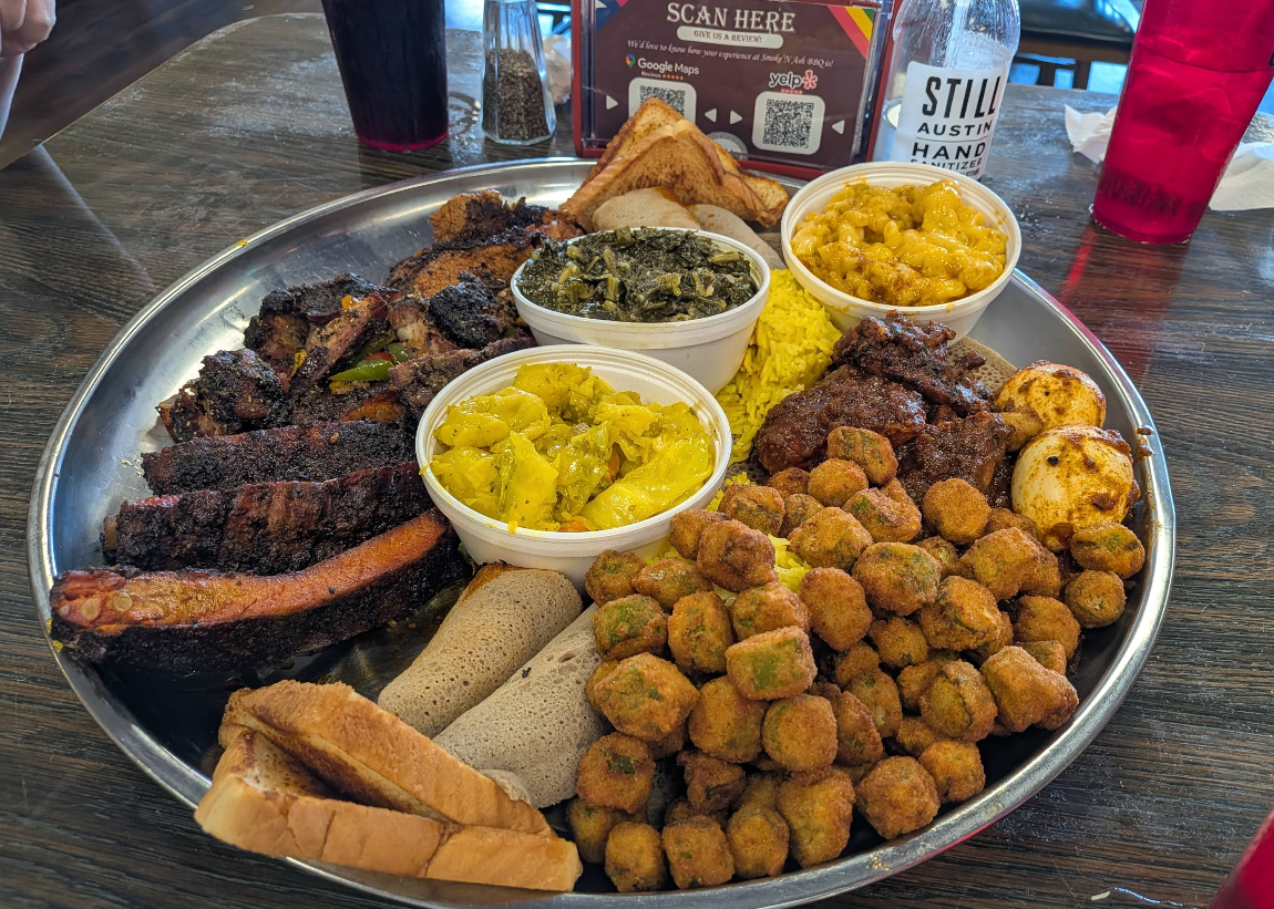 A large metal platter filled with assorted barbecue meats, including some of the best ribs in America, plus cornbread, toast, collard greens, macaroni and cheese, yellow curried vegetables, fried okra, and deviled eggs on a wooden table with drinks in the background.
