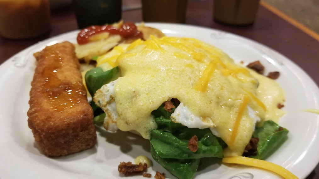 A cheap all you can eat breakfast plate featuring a hash brown topped with syrup, eggs over spinach covered in melted cheese sauce, and a potato round with ketchup, all served on a white plate.