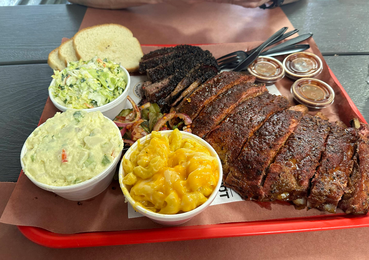 A tray of barbecue food featuring some of the best ribs in America, savory burnt ends, three sides (cole slaw, potato salad, macaroni and cheese), pickles, cups of barbecue sauce, and slices of white bread on brown paper.