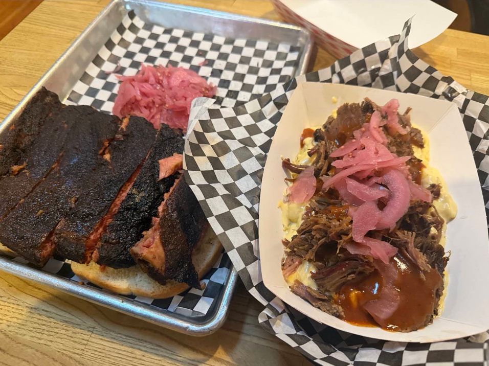A tray with the best ribs in America on toast and pickled onions beside macaroni and cheese topped with pulled beef, barbecue sauce, and pickled onions, all served on a wooden table.