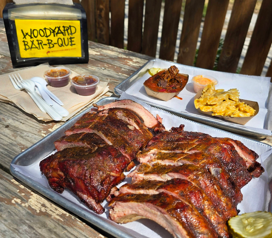 A tray of smoked ribs—hailed as some of the best ribs in America—is served with macaroni and cheese, baked beans, pickles, and barbecue sauce on a rustic wooden table beside a napkin holder labeled “Woodyard Bar-B-Que.”