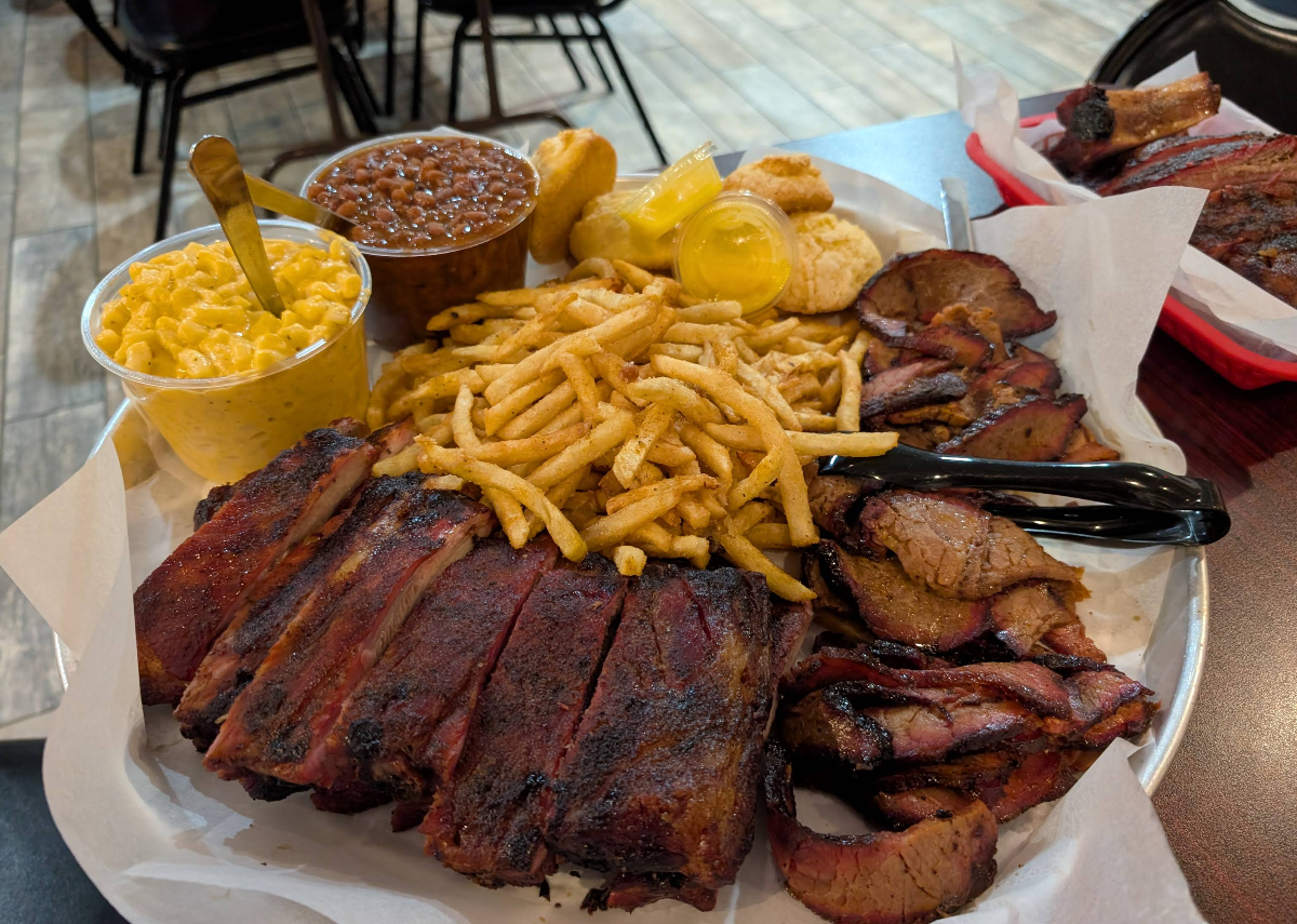 A platter of barbecue food featuring the best ribs in America, brisket slices, French fries, macaroni and cheese, baked beans, cornbread, a biscuit, and two cups of yellow dipping sauce on a table in a restaurant.