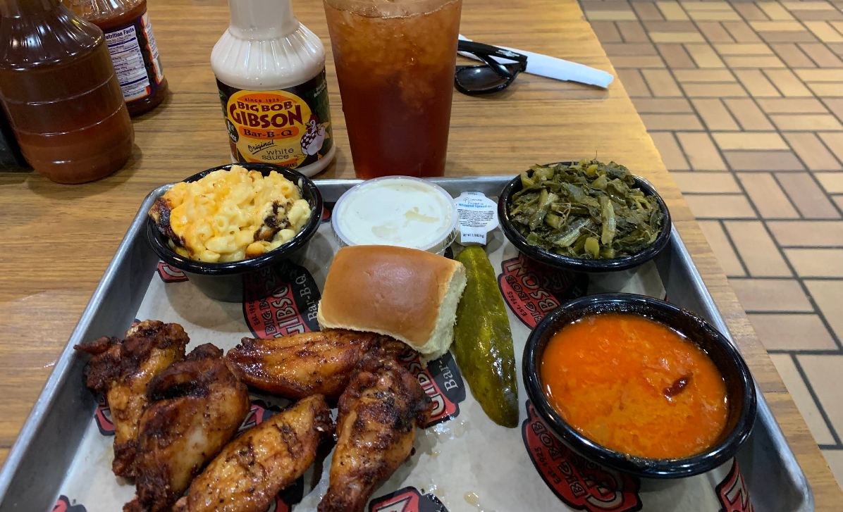 A tray of barbecue food with smoked chicken wings, macaroni and cheese, collard greens, a roll, a pickle spear, dipping sauces, and a bowl of red soup—plus iced tea and the best ribs in America sauce on the side.