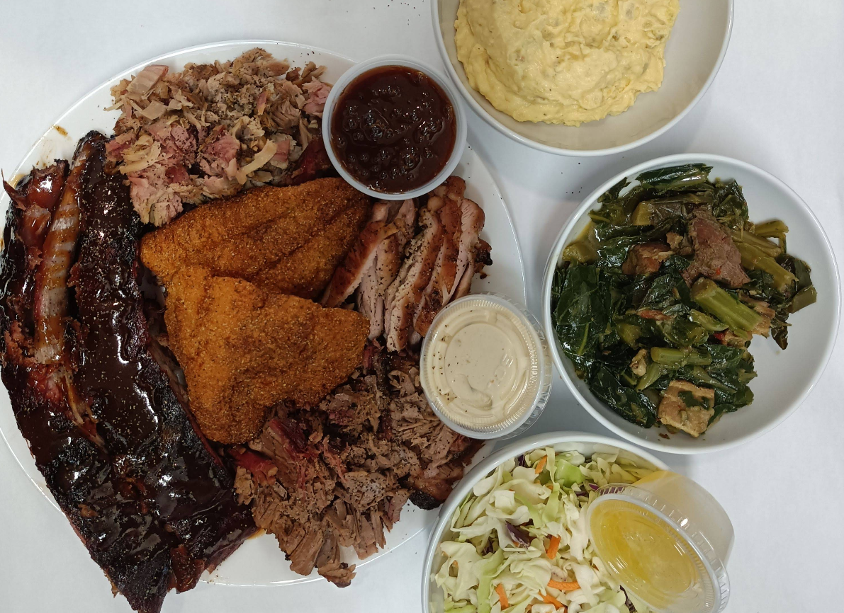 A plate of assorted barbecue meats, featuring the best ribs in America alongside fried catfish, pulled pork, and smoked chicken, served with mashed potatoes, collard greens, coleslaw, barbecue sauce, and dressing.