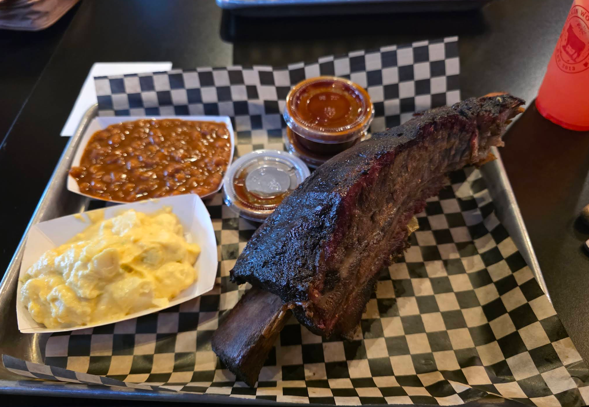 A tray with a large smoked beef rib—truly some of the best ribs in America—served alongside baked beans, macaroni and cheese, barbecue sauce on black-and-white checkered paper, and a pink drink in the background.