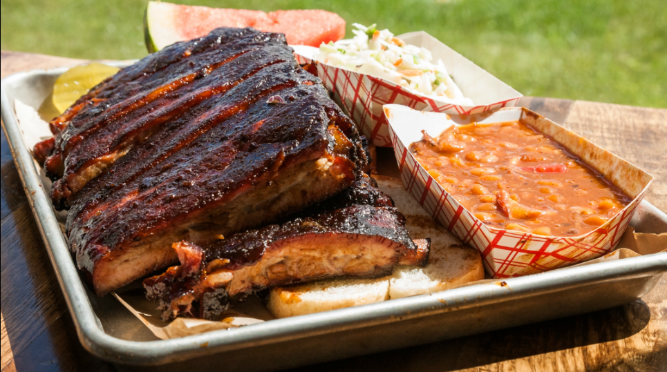 A tray of the best ribs in America served with pickles, a slice of watermelon, coleslaw, and baked beans in paper trays, all set on a wooden table with grass visible in the background.