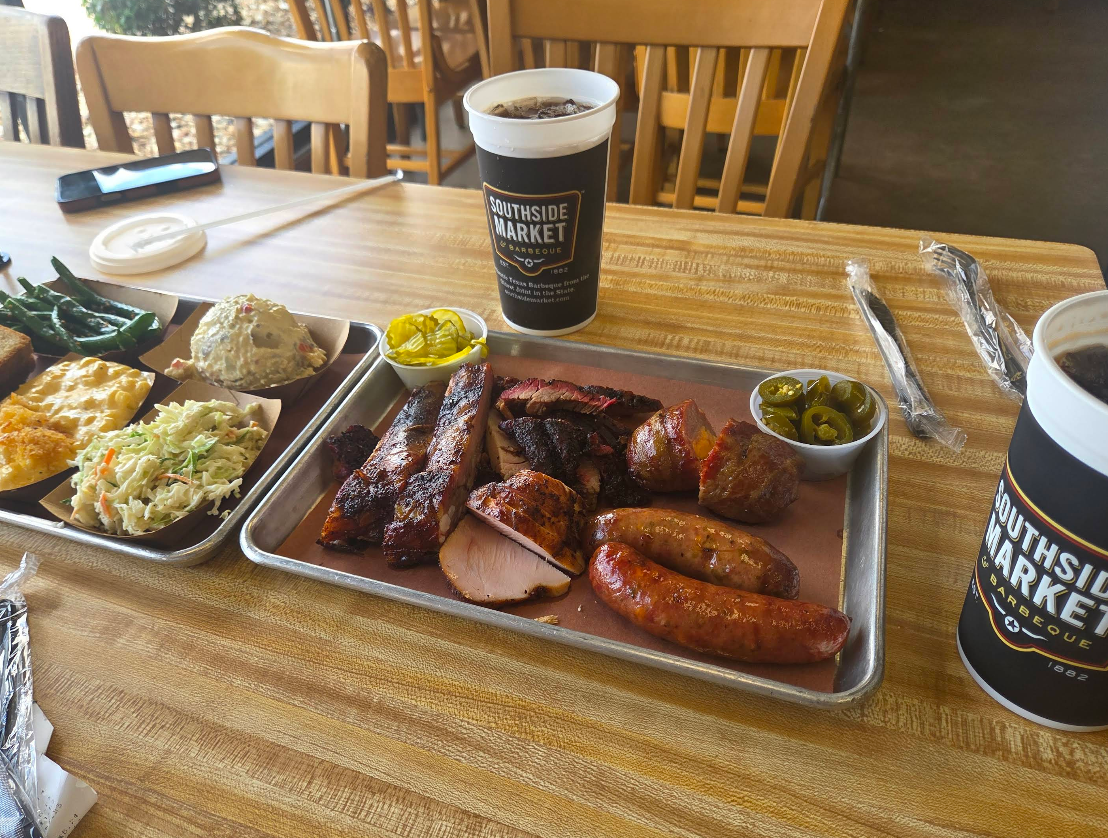 A tray with assorted barbecue meats, including some of the best sausages in America, pickles, and jalapeños sits on a wooden table alongside cups with “Southside Market” logo and plates of classic sides like coleslaw, potato salad, and green beans.