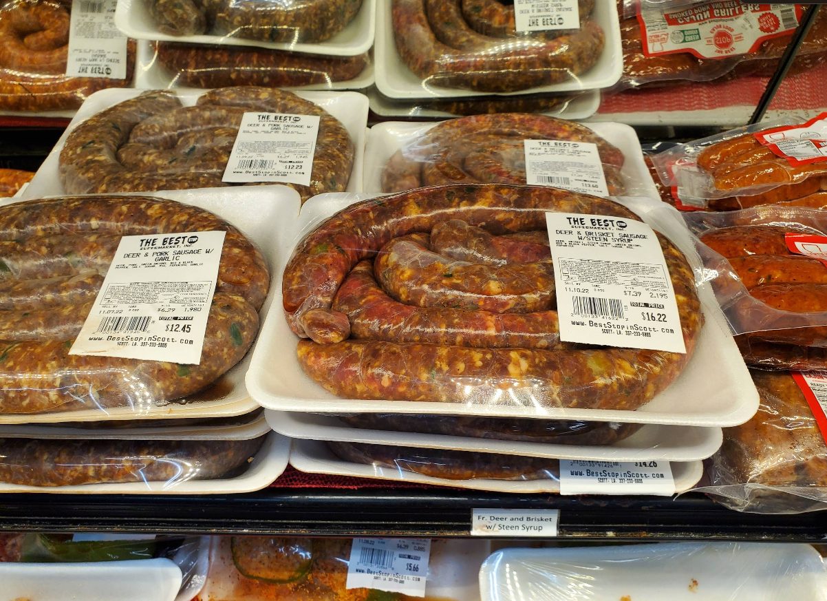 Plastic-wrapped trays of raw beef and pork sausages—touted among the best sausages in America—are displayed on a grocery store shelf, with white labels listing product information and prices. Other packaged meats are visible nearby.