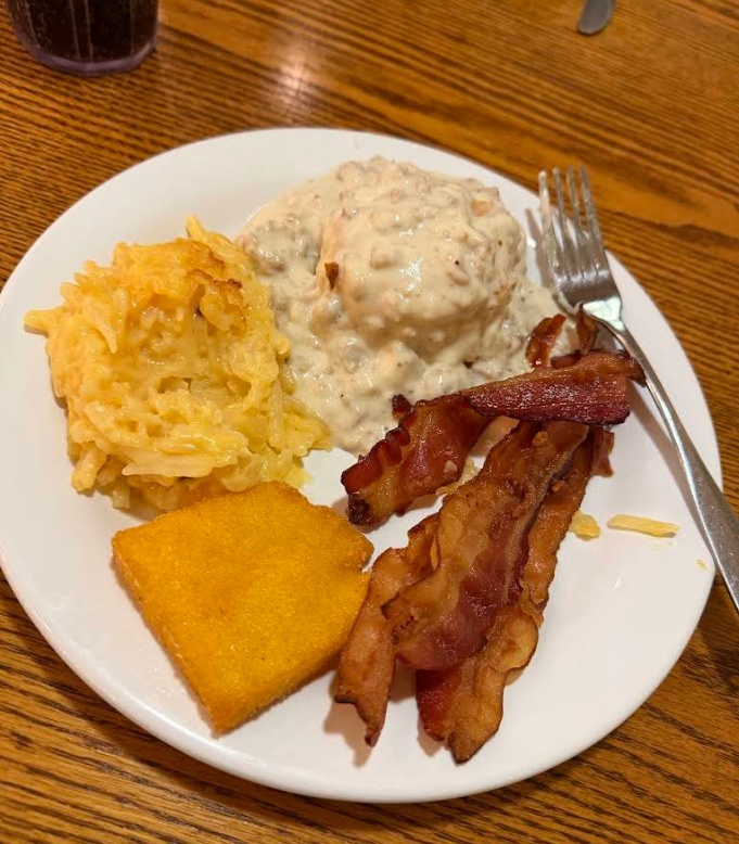A plate from a cheap all you can eat breakfast, featuring crispy bacon strips, a square of cornbread, hash brown casserole, and a biscuit topped with white sausage gravy. A fork rests on the side atop the wooden table.
