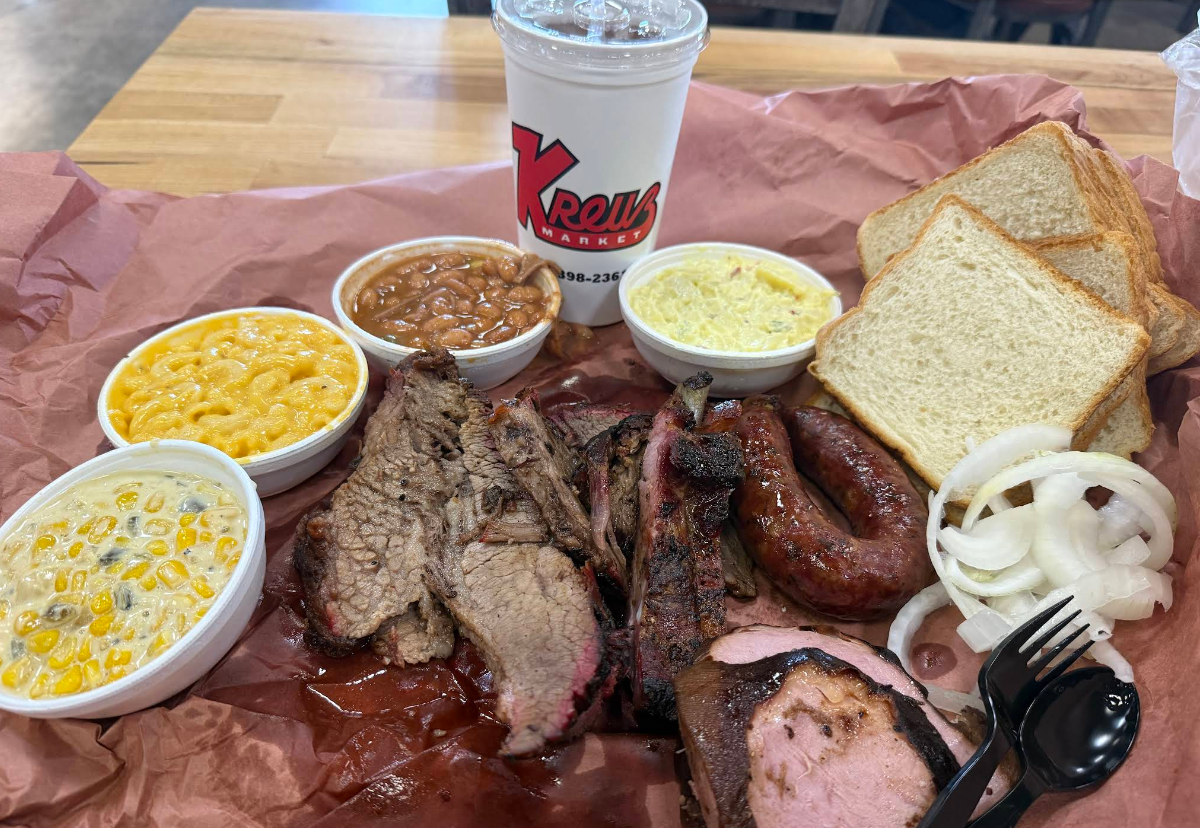 A BBQ platter with sliced brisket, the best sausages in America, ribs, beans, creamed corn, macaroni and cheese, potato salad, sliced onions, white bread, a plastic fork, and a drink cup on brown butcher paper.