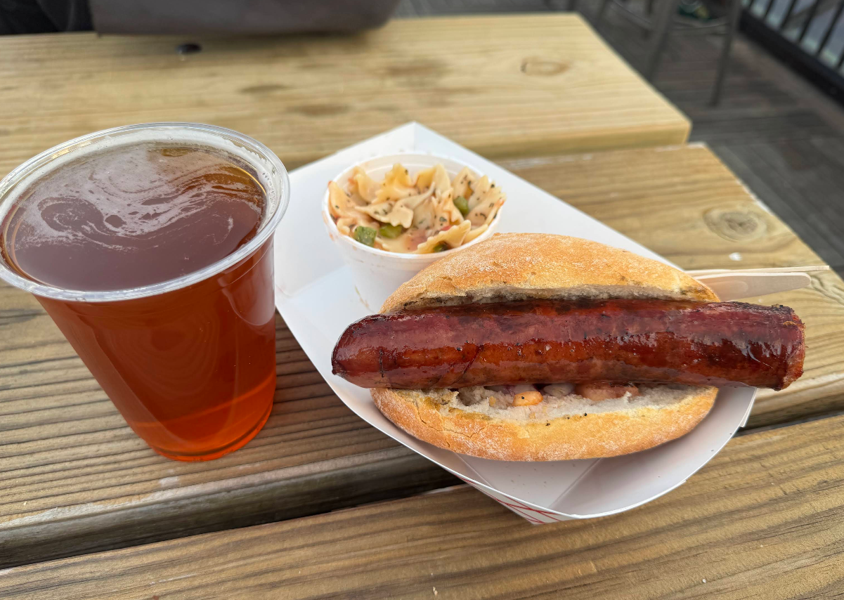 Enjoy one of the best sausages in America served on a sandwich bun, paired with pasta salad and a refreshing cup of amber-colored beer atop a wooden picnic table.