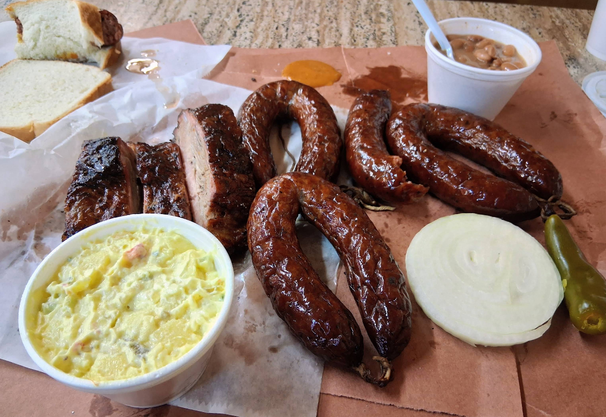 A barbecue meal featuring some of the best sausages in America, smoked sausage links, pork ribs, sliced white bread, potato salad, pinto beans, sliced onions, a whole jalapeño, and a dollop of barbecue sauce served on butcher paper.