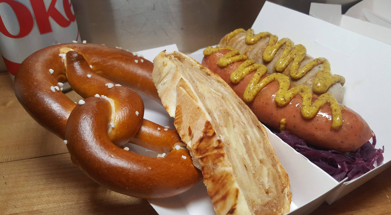 A soft pretzel, a slice of layered pastry, and one of the best sausages in America topped with mustard served over red cabbage in a takeout tray. A Diet Coke can is partially visible in the background.