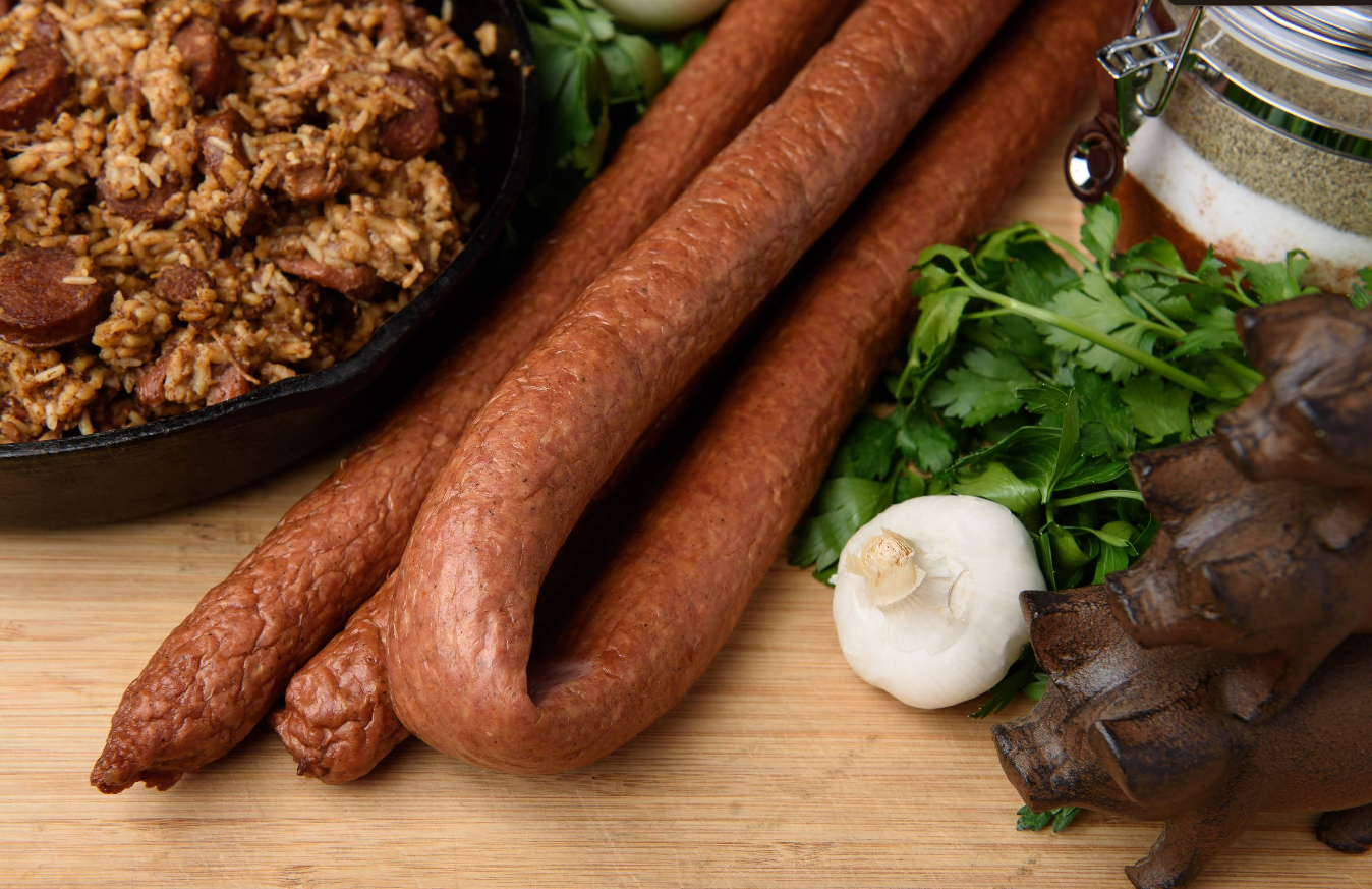 Two smoked sausages, known among the best sausages in America, rest on a wooden board near fresh cilantro, a head of garlic, seasoning jar, and a cast-iron skillet filled with savory sausage rice beside a cast-iron trivet.
