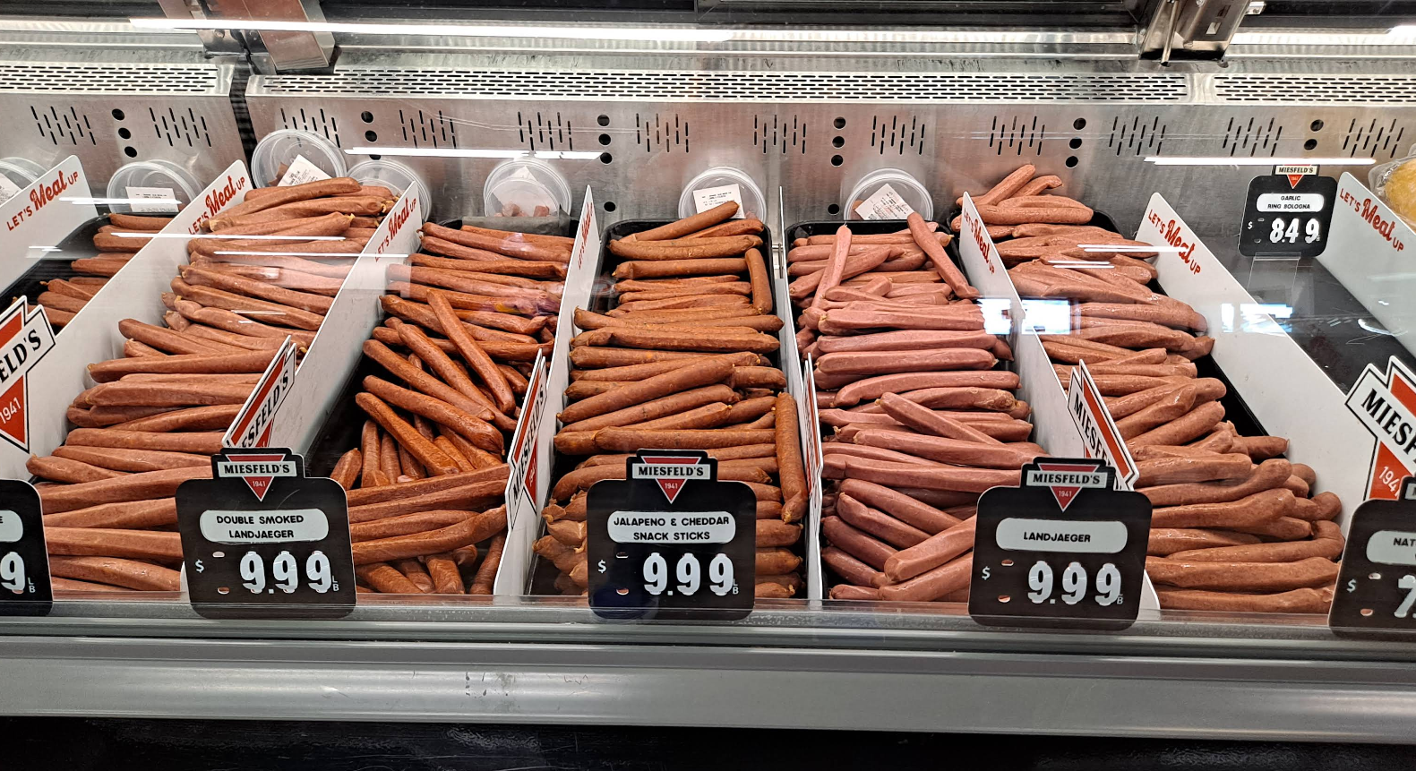 A grocery store meat counter displays five sections of the best sausages in America, including double smoked, jalapeno cheddar, and landjaeger, each labeled with price tags reading $9.99 per pound.