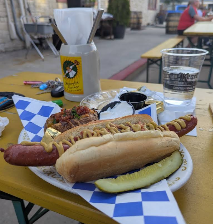 A plate featuring one of the best sausages in America as a hot dog topped with mustard, alongside a pickle spear and a baked potato with chili and sour cream, sits on a yellow picnic table with drinks and napkins nearby.