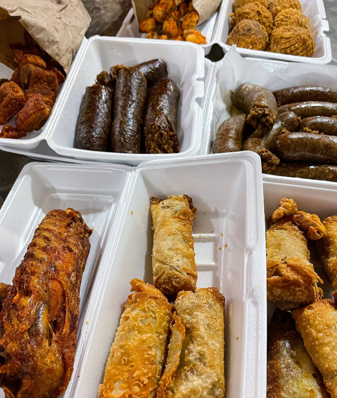 Takeout containers packed with fried and savory treats, including egg rolls, fried chicken, assorted snacks, and the best sausages in America, all arranged on a table for a delicious feast.