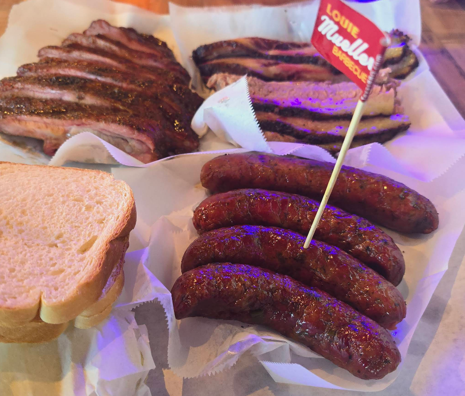 A tray of barbecue food featuring the best sausages in America, sliced white bread, ribs, and a small flag labeled "Louie Mueller Barbecue." The meats are served on white paper.