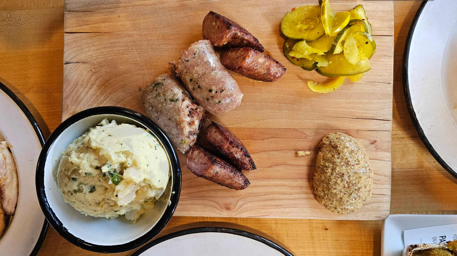 A wooden board with some of the best sausages in America, a pile of yellow pickles, a scoop of grainy mustard, and a bowl of mashed potatoes garnished with herbs on a wooden table.