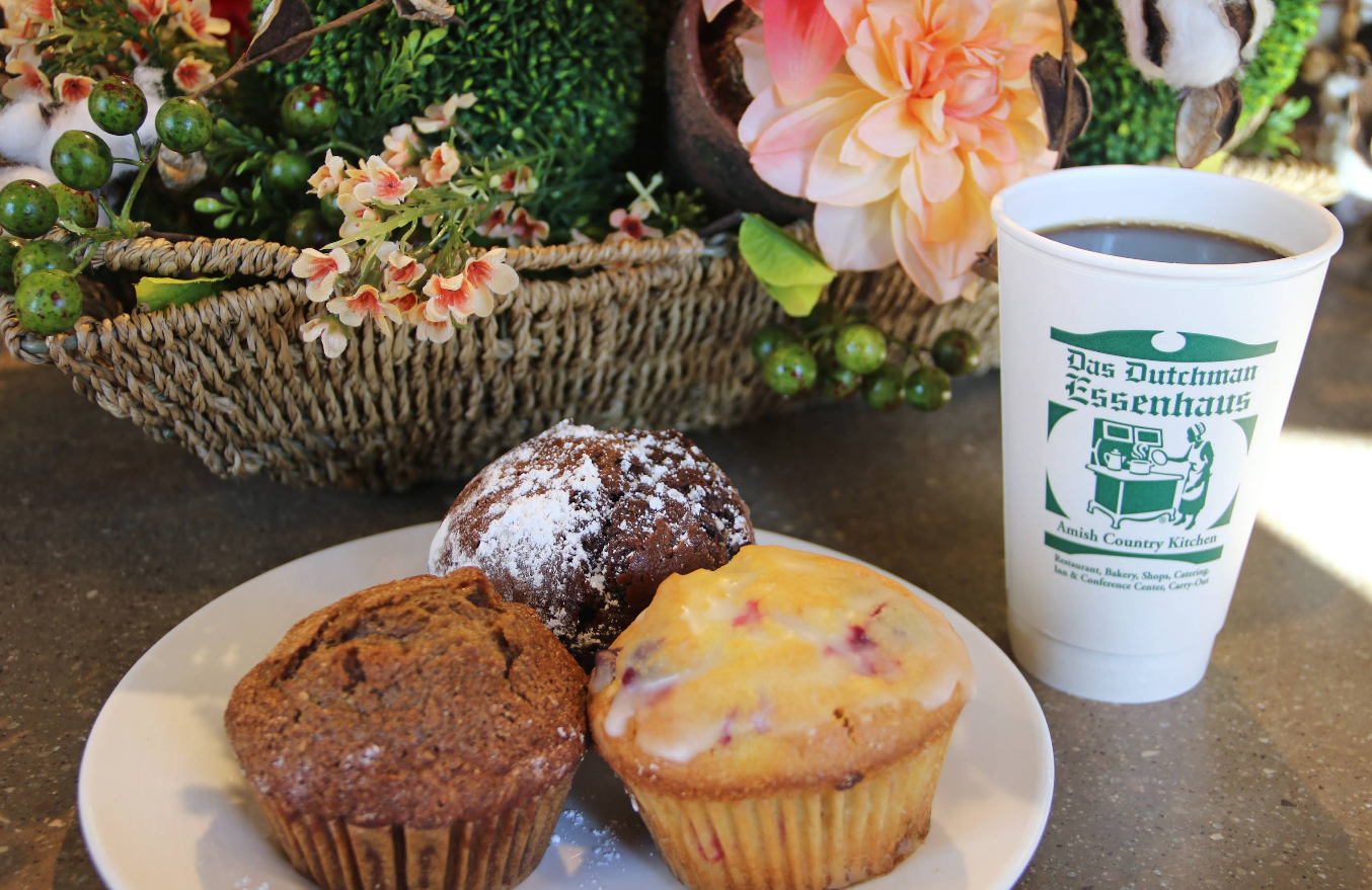 Three muffins sit on a white plate next to a cup of coffee in a white to-go cup labeled "Das Dutchman Essenhaus," hinting at the delightful, cheap all you can eat breakfast experience with flowers and greenery in the background.