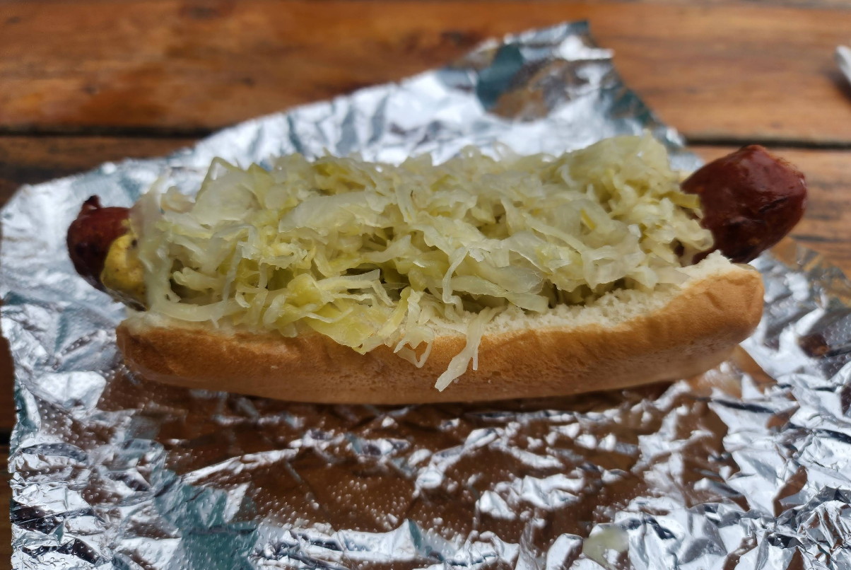 A grilled hot dog—one of the best sausages in America—in a bun topped with sauerkraut, placed on crumpled aluminum foil on a wooden table.