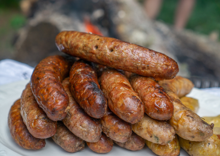 A close-up of a pile of grilled sausages, hailed as some of the best sausages in America, stacked on a white plate with a fire pit and blurred outdoor background visible behind them.