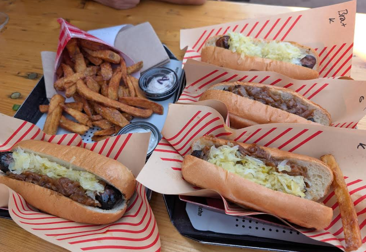 Four hot dogs, featuring some of the best sausages in America, are topped with sauerkraut and served on striped paper next to golden French fries and dipping sauces. A person's hand is partially visible in the background.