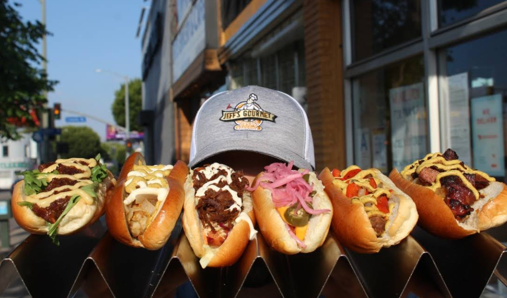 A person wearing a Jeff’s Gourmet cap is partially hidden behind a row of six gourmet hot dogs—topped with colorful ingredients and possibly featuring the best sausages in America—displayed outdoors in front of a building.