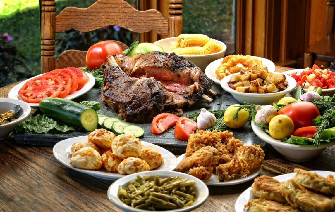 A wooden table set with assorted Southern-style foods, including roast meat, fried chicken, sliced tomatoes, corn on the cob, green beans, biscuits, fried potatoes, fresh vegetables, and bowls of salad—perfect for a cheap all you can eat breakfast.