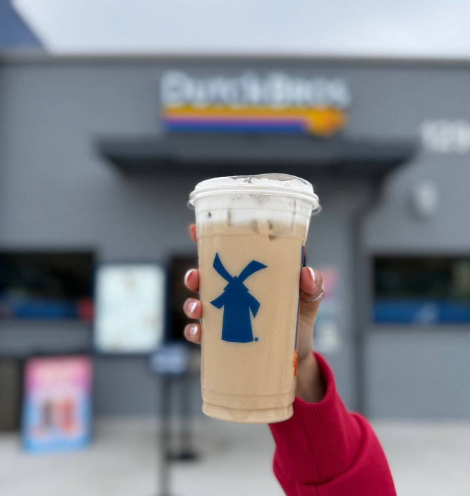 A hand holding an iced coffee drink in a clear cup with a blue windmill logo, in front of a Dutch Bros coffee shop. The background is blurred, focusing on the drink and hand with pink-painted nails.