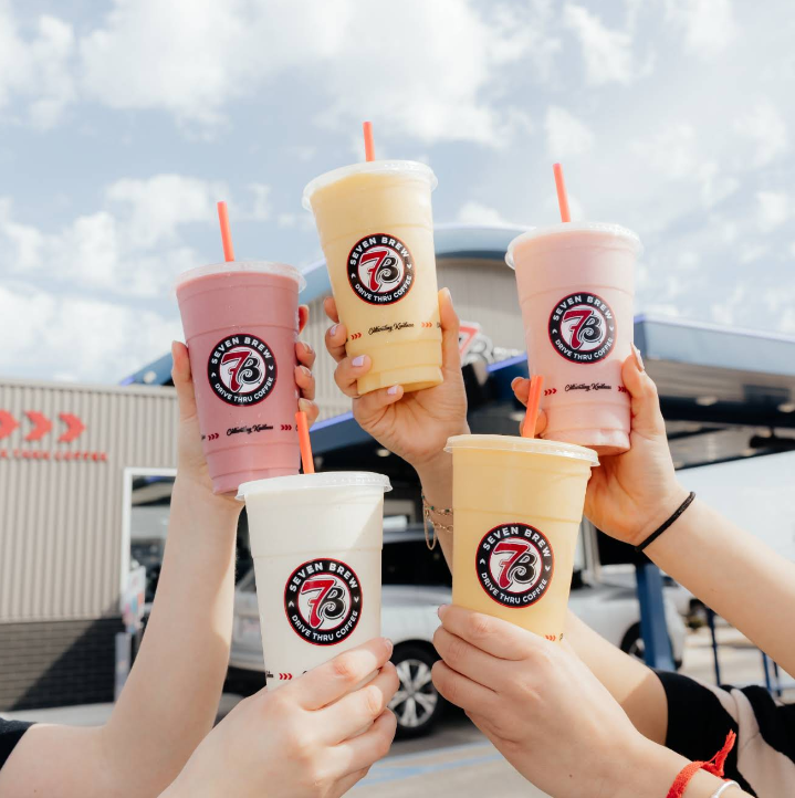 Five hands hold colorful drinks in clear cups with red straws, each featuring the 7 Brew logo. The drinks are raised together outside, with a building and a partly cloudy sky in the background.