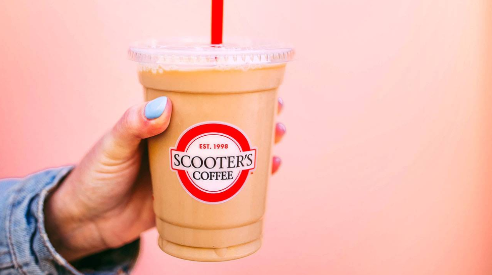 A hand with light blue nail polish holds an iced coffee in a plastic cup with a red straw and a Scooter’s Coffee logo against a pink background.