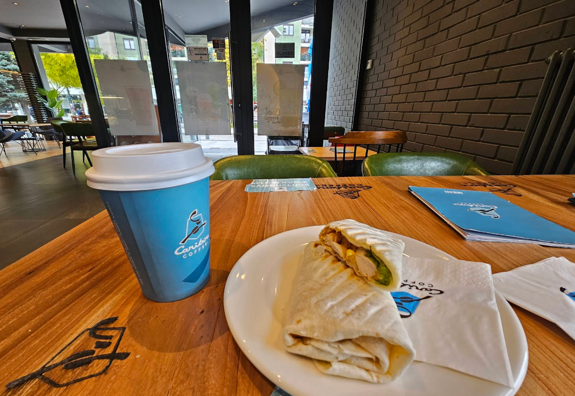 A wrap sandwich cut in half on a plate with a napkin, next to a blue takeaway coffee cup, on a wooden table in a modern, well-lit café with large windows and green chairs.