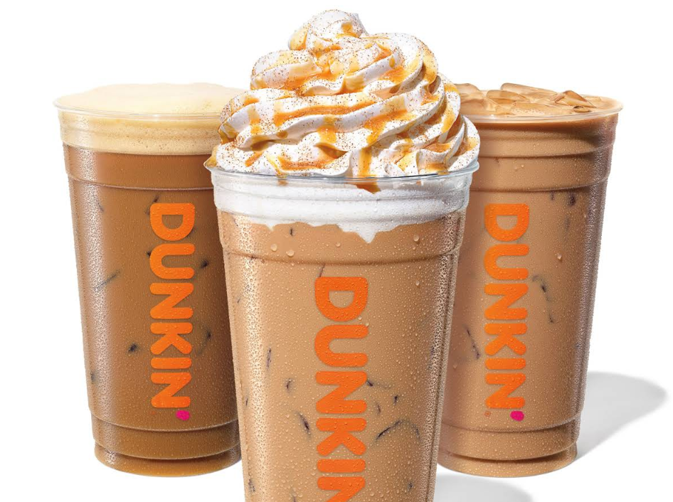 Three Dunkin’ iced coffee drinks in clear cups with orange “DUNKIN’” logo; one topped with whipped cream and caramel drizzle, and two plain iced coffees, one slightly frothy and the other with visible ice cubes.